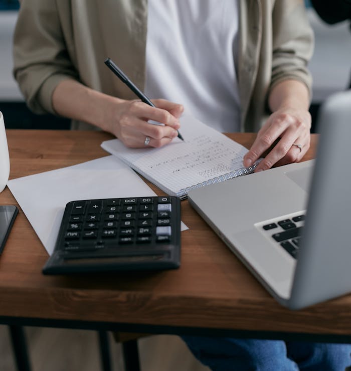 pexels-photo-6962993 A woman writes financial calculations in a notebook, using a calculator and laptop at a wooden desk.